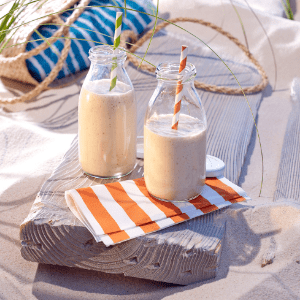 Bananen-Kokos-Shake in kleinen Glasflaschen auf einer Servierte stehend auf einem Holzbrett im Sand mit einer Tasche im Hintergrund