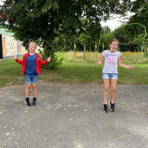 Kleines Mädchen und kleiner Junge springen auf einer asphaltierten Fläche vor einer Wiese Springseil