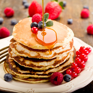 Pancakes mit Sirup und Beeren zusammen mit einer Gabel auf einem Teller, im Hintergrund Beeren auf einem Holztisch